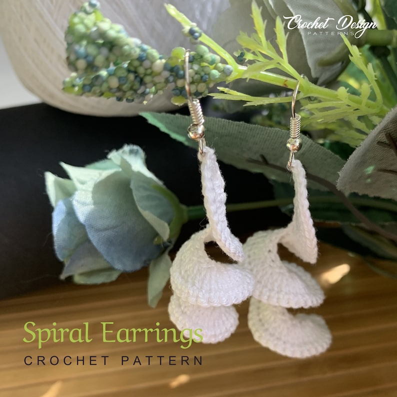 K&ouml;nnte beinhalten: Wei&szlig;e geh&auml;kelte Spiralohrringe mit silbernen Haken. Die Ohrringe werden vor einem Hintergrund aus gr&uuml;nem Laub gezeigt. "Spiral Earrings CROCHET PATTERN" ist in gr&uuml;nem Text am unteren Rand des Bildes geschrieben.