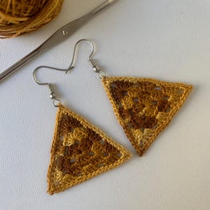 May include: Handmade crochet earrings in a triangular shape, featuring shades of brown and gold. The earrings have silver-colored hooks and a detailed, openwork design. A crochet hook and yarn are visible in the background.