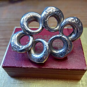 Könnte beinhalten: Eine silberne Brosche mit floralem Design, bestehend aus sechs ineinandergreifenden Kreisen. Die Brosche befindet sich in einer roten Schachtel.