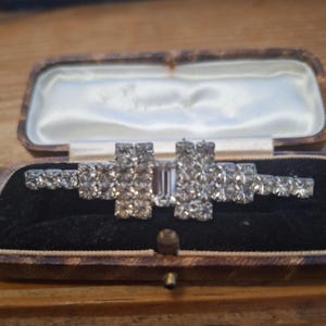 Pode incluir: Um broche de barra em tom prateado com uma pedra central transparente de corte esmeralda, ladeada por dois grupos retangulares de strass transparentes, com uma fila de strass menores estendendo-se em cada lado do alfinete.