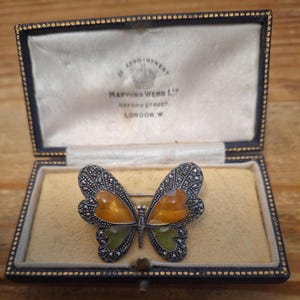 May include: A butterfly brooch featuring amber and green translucent stones set in silver-toned metal with marcasite accents. The brooch is displayed inside a navy blue rectangular jewelry box with white satin lining and the text Mappin & Webb Ltd.