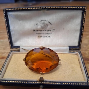 Puede incluir: Un broche ovalado de piedra preciosa facetada color ámbar montado en un marco de metal dorado. El broche se exhibe dentro de una caja de presentación rectangular azul marino con un interior color crema con el texto Mappin & Webb Ltd.