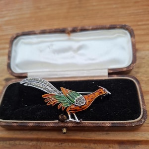Puede incluir: Un broche de ave en tono plateado con detalles de esmalte naranja y verde vibrante en el cuerpo y las alas, acentuado con pequeñas piedras de marcasita brillantes a lo largo de las largas plumas de la cola y el ala, presentado en un joyero rectangular.