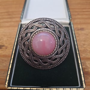 May include: A round brooch featuring a central smooth pink cabochon stone set within a dark silver-toned metal frame. The frame displays an intricate, raised braided or woven pattern that encircles the stone, creating a textured border.