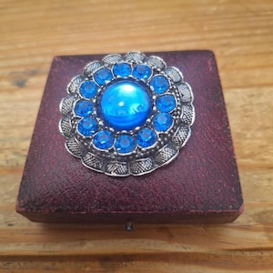 Op de afbeelding: Een ronde broche met een grote, gladde, felblauwe centrale cabochon omringd door een ring van kleinere, gefacetteerde blauwe kristallen. De zetting is gemaakt van getextureerd, antiek zilverkleurig metaal met een geschulpte, decoratieve rand.