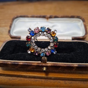 Puede incluir: Un broche circular con una doble hilera de pedrería transparente y un anillo exterior de piedras facetadas multicolores en tonos azul, rojo, verde, naranja y rosa, montado en un marco metálico y presentado en un joyero rectangular.
