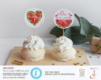 Peut inclure: Deux cupcakes avec glaçage blanc et des décorations sur le thème de la pastèque. L'une est en forme de cœur avec le texte "Alexandra is 1", l'autre dit "One in a melon". Les cupcakes sont sur une planche en bois.