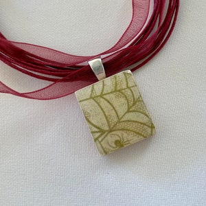 May include: A square pendant necklace with a cream and olive green spiderweb design. The pendant is attached to a silver bail and strung on multiple strands of burgundy sheer ribbon.