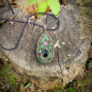 May include: A handcrafted pendant necklace featuring a teardrop-shaped pendant with a black stone, a purple stone, and green leaf accents. The pendant hangs from a black cord with a bronze clasp. The text "Wolf Spirit Arts by Sara" is visible.