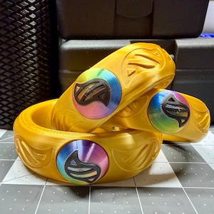 Puede incluir: Tres pulseras doradas impresas en 3D. Cada pulsera presenta un emblema circular multicolor con un diseño negro estilizado. Las pulseras están apiladas y dispuestas para mostrar su diseño y textura. El fondo es gris neutro.