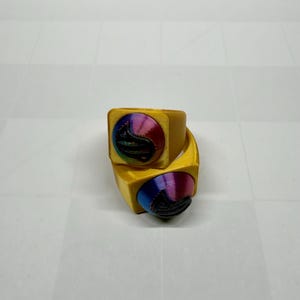 Puede incluir: Dos anillos dorados con caras cuadradas. Cada anillo presenta un diseño circular con un degradado arcoíris y un elemento negro texturizado. Los anillos están apilados, mostrando el diseño y las variaciones de color.