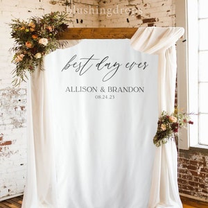 May include: White backdrop with the text "best day ever" and the names "Allison & Brandon" and the date "08.24.23" written in black calligraphy. The backdrop is hung on a wooden frame and decorated with white fabric drapes and floral arrangements.