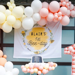 May include: A pink, yellow, and white balloon arch with a banner that reads "Blair's 1st Bee-day" and a large pink number one balloon.