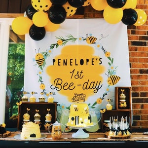 May include: A bee-themed first birthday party table setting with a yellow and black cake, cupcakes, and other treats. The backdrop features a white banner with the text "PENELOPE'S 1st Bee-day" in black lettering.