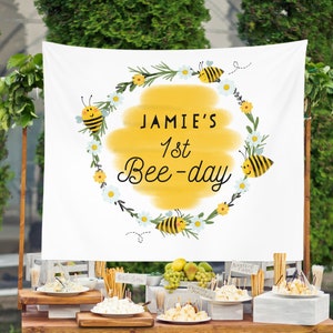 May include: A white banner with a yellow circle and the text "Jamie's 1st Bee-day" surrounded by bees and flowers. The banner is hanging from a wooden stand with a table in front of it. The table is decorated with food and drinks for a party.