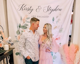 Puede incluir: Una pareja posa para una foto frente a un telón de fondo floral. El telón de fondo tiene el texto "Kirsty & Stephen are engaged". La mujer lleva un vestido rosa y el hombre lleva una camisa blanca abotonada y pantalones vaqueros azules.