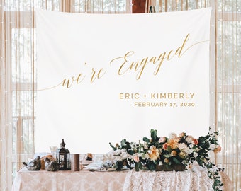 May include: White backdrop with gold calligraphy that reads "we're engaged" with the names Eric + Kimberly and the date February 17, 2020. The backdrop is displayed on a table with a blush pink tablecloth and white lace overlay. There are floral arrangements on the table with peach, white, and yellow flowers.