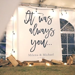 May include: A white sign with black calligraphy that reads "It was always you..." with the names Milena & Michael and the date 10.30.2021 below. The sign is hanging in a white tent with string lights.