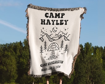May include: White woven throw blanket with black text and illustrations. The text reads "Camp Hayley Camp Bachelorette Bass Lake CA 2024". The illustration is of a mountain range with a tent, trees, a campfire, and stars.