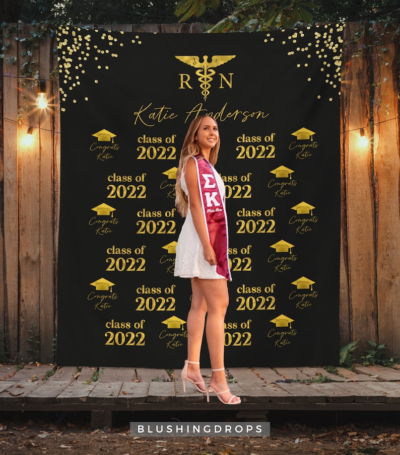 May include: A black graduation backdrop with gold confetti and the text "RN" and "Katie Anderson" in gold. The backdrop also features the text "class of 2022" repeated multiple times. A woman is standing in front of the backdrop wearing a white dress and a maroon graduation stole.