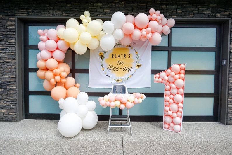 May include: A pink, yellow, and white balloon arch with a banner that reads "Blair's 1st Bee-day" and a large pink number one balloon.