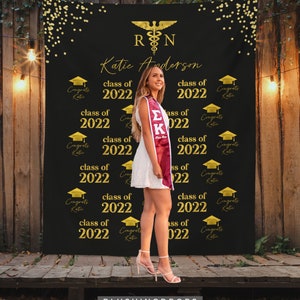 May include: A black graduation backdrop with gold confetti and the text "RN" and "Katie Anderson" in gold. The backdrop also features the text "class of 2022" repeated multiple times. A woman is standing in front of the backdrop wearing a white dress and a maroon graduation stole.
