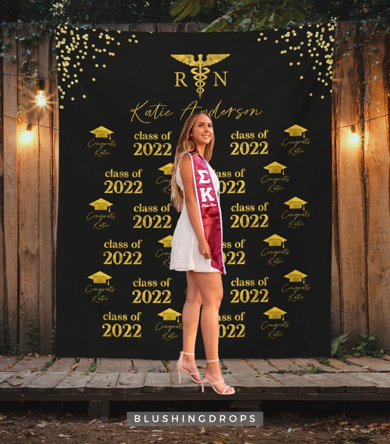 May include: A black graduation backdrop with gold confetti and the text "RN" and "Katie Anderson" in gold. The backdrop also features the text "class of 2022" repeated multiple times. A woman is standing in front of the backdrop wearing a white dress and a maroon graduation stole.
