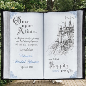 May include: A white book with a blue and gray design on the cover. The book is open to a page with a drawing of a castle and the text "Once upon a time... In a kingdom not so far, far away there lived a beautiful princess who said "YES" to her prince... Let's celebrate Venessa's Bridal Shower July 16th, 2021...and she lived Happily Ever After."