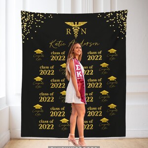 May include: A black graduation backdrop with gold confetti and the text "RN" and "Katie Anderson" in gold script. The backdrop also features the text "class of 2022" repeated multiple times and gold graduation caps.