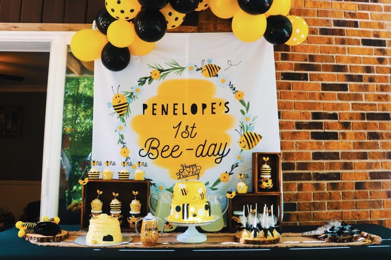 May include: A bee-themed first birthday party table setting with a yellow and black cake, cupcakes, and other treats. The backdrop features a white banner with the text "PENELOPE'S 1st Bee-day" in black lettering.