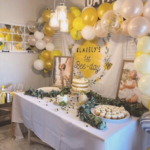 May include: A white tablecloth with a yellow and white balloon arch, a white cake with yellow frosting, and a banner that says "Blakely's 1st Bee-day".