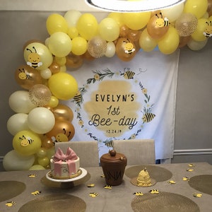 May include: A yellow and gold balloon arch with bee decorations for a first birthday party. The banner says "Evelyn's 1st Bee-day 12.24.19". There is a cake with a pink bow and a honey pot on a table with bee-themed decorations.