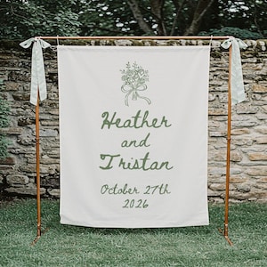 May include: A white fabric wedding backdrop with the names "Heather and Tristan" and the date "October 27th 2026" in green script. The backdrop is supported by a copper-colored metal frame and adorned with green ribbons.