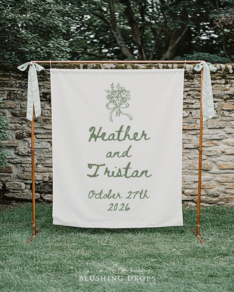 May include: A white fabric wedding backdrop with the names "Heather and Tristan" and the date "October 27th 2026" in green script. The backdrop is supported by a copper-colored metal frame and adorned with green ribbons.