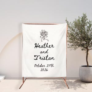 May include: A white fabric wedding sign with the names "Heather and Tristan" and the date "October 27th 2026" in black script. The sign is supported by a copper-colored metal frame, with a floral illustration at the top.