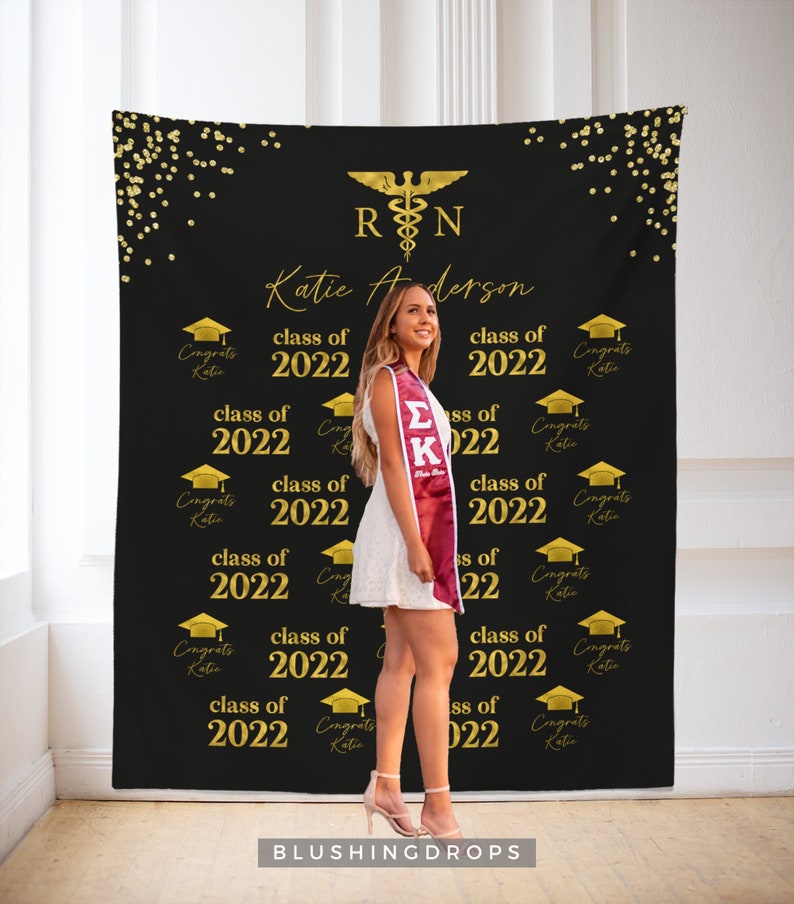 May include: A black backdrop with gold confetti and the text "RN" in a gold medical symbol. The text "Katie Anderson" is written in gold lettering. The backdrop is decorated with gold graduation caps and the text "class of 2022" repeated multiple times.