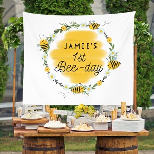 May include: A white banner with a yellow circle and a wreath of flowers and bees. The text on the banner reads "Jamie's 1st Bee-day".