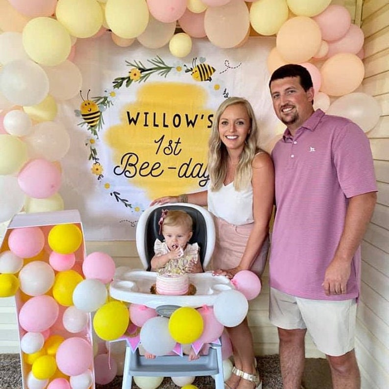 May include: A yellow and white banner with the text "Willow's 1st Bee-day" and a bee illustration. A baby girl sits in a high chair decorated with yellow, pink, and white balloons. A woman and a man stand behind the baby, smiling.