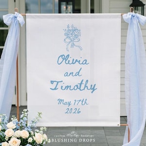 May include: A white wedding sign with the names "Olivia and Timothy" in light blue script, along with the date "May 17th 2026." The sign is adorned with a floral illustration and light blue fabric. The sign is supported by a copper-colored frame.