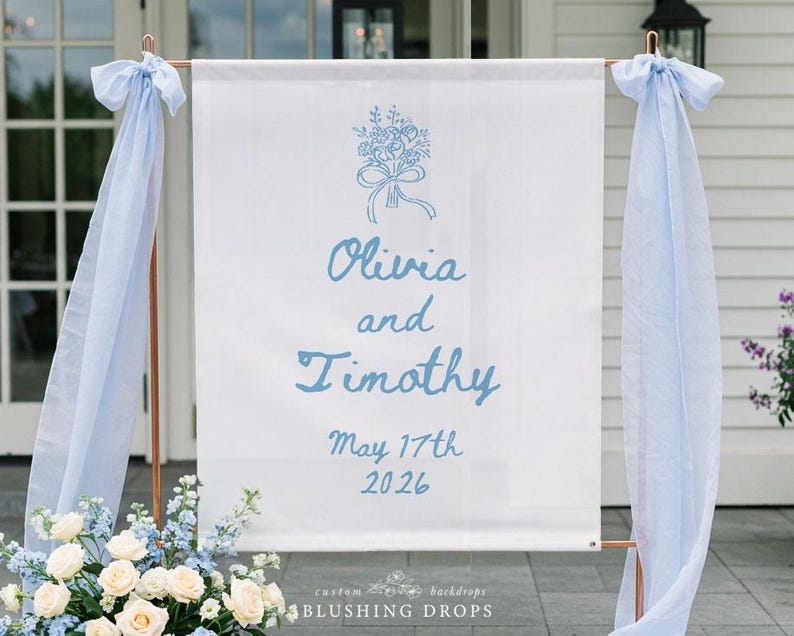 May include: A white wedding sign with the names "Olivia and Timothy" in light blue script, along with the date "May 17th 2026." The sign is adorned with a floral illustration and light blue fabric. The sign is supported by a copper-colored frame.