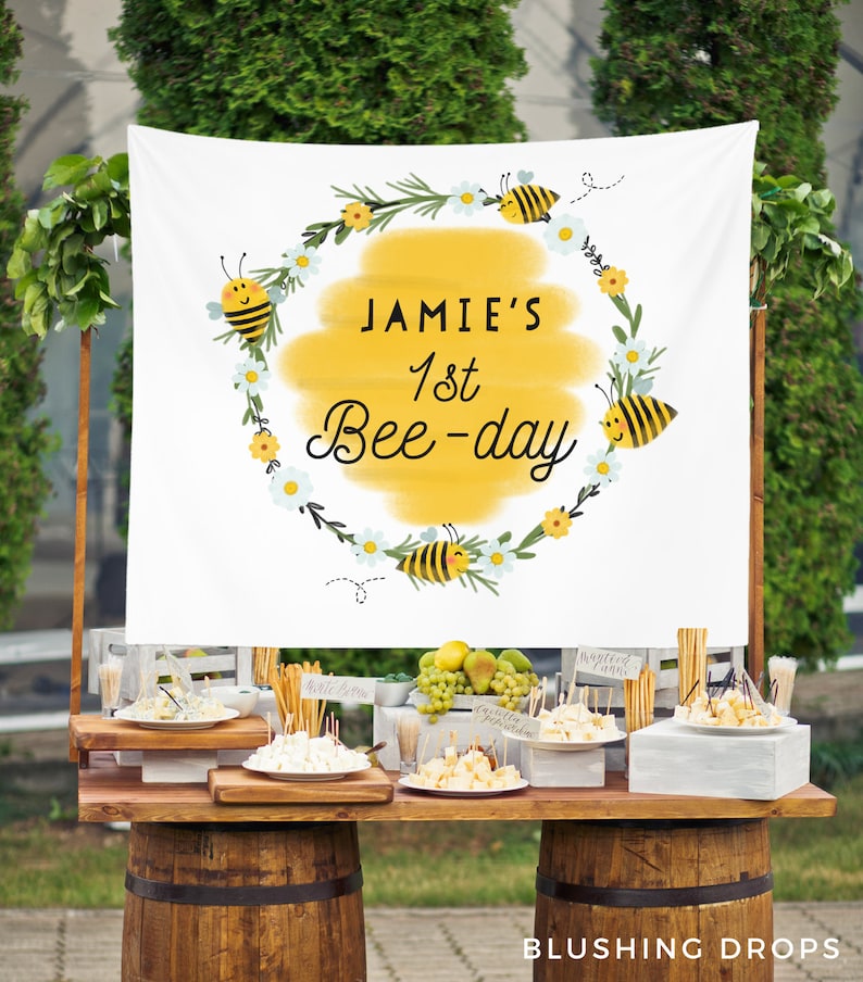 May include: A white banner with a yellow circle and the text "Jamie's 1st Bee-day" surrounded by bees and flowers. The banner is hanging from a wooden stand with a table in front of it. The table is decorated with food and drinks for a party.