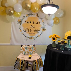May include: A white banner with a yellow wreath and bees, reading "Gabrielle's 1st Bee-day 9.30.2019". A high chair with a yellow and black bee-themed fabric and a yellow and black banner with the number "1" and a bee. A black tablecloth with a bee-themed cake and cupcakes.
