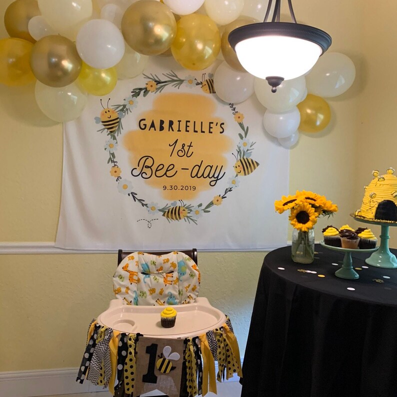 May include: A white banner with a yellow wreath and bees, reading "Gabrielle's 1st Bee-day 9.30.2019". A high chair with a yellow and black bee-themed fabric and a yellow and black banner with the number "1" and a bee. A black tablecloth with a bee-themed cake and cupcakes.