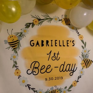 May include: A white banner with a yellow and green wreath with white flowers and bees. The banner says "Gabrielle's 1st Bee-day 9.30.2019".