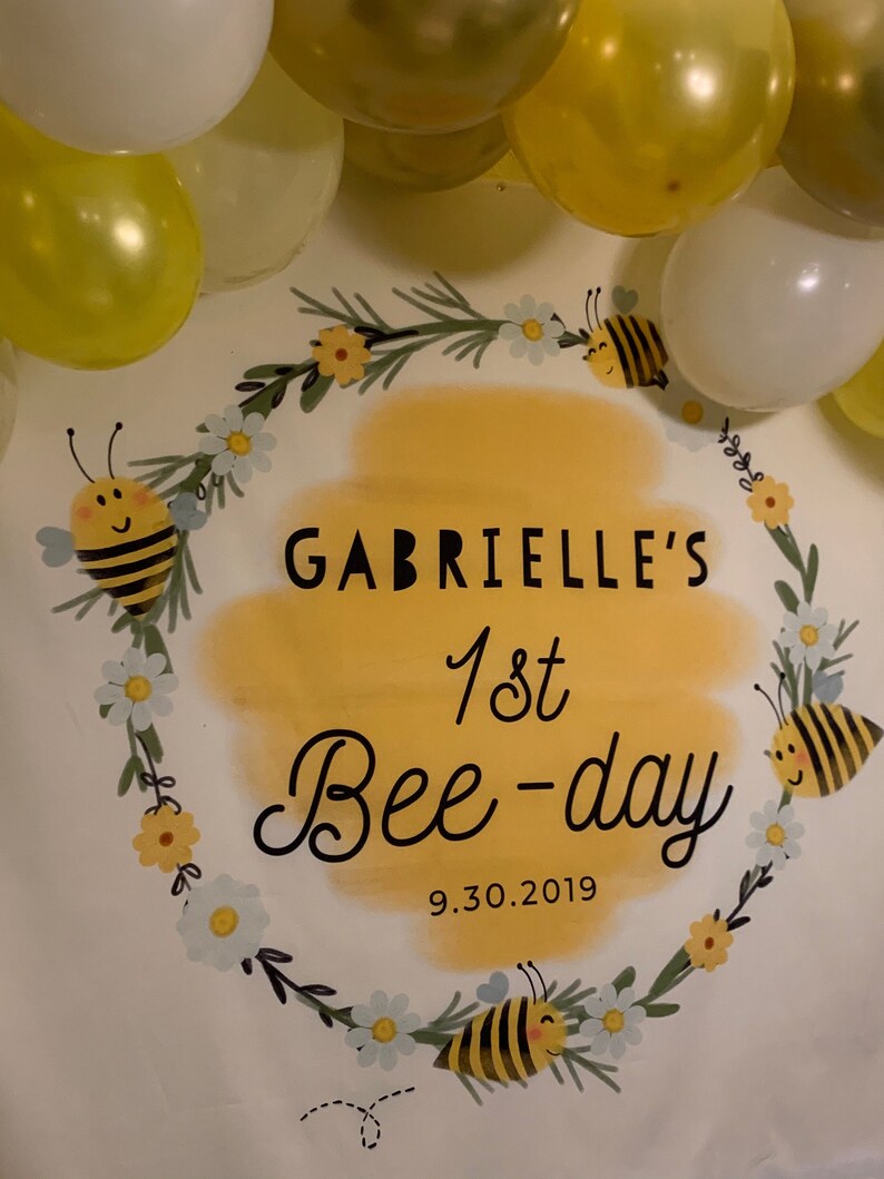 May include: A white banner with a yellow and green wreath with white flowers and bees. The banner says "Gabrielle's 1st Bee-day 9.30.2019".