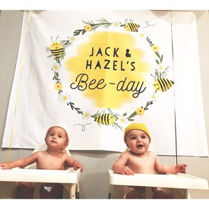 May include: Two babies wearing hats sit in high chairs in front of a white banner with a yellow circle and the text "Jack & Hazel's Bee-day" in black lettering. The circle is surrounded by a wreath of green leaves, yellow flowers, and bees.