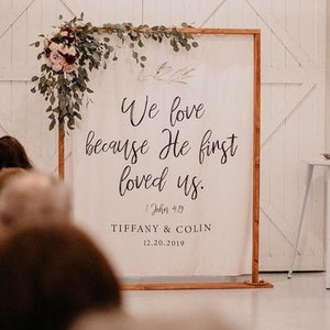 May include: A white canvas sign with a wooden frame, decorated with greenery and flowers, reads "We love because He first loved us."  The sign also includes the scripture reference "1 John 4:19" and the names "Tiffany & Colin" with the date "12.20.2019".