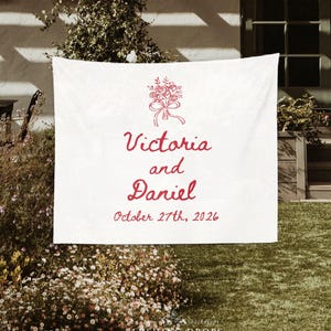 May include: White wedding backdrop with red floral bouquet illustration, the names "Victoria and Daniel", and the date "October 27th, 2026". The backdrop is hanging outdoors with greenery in the background.