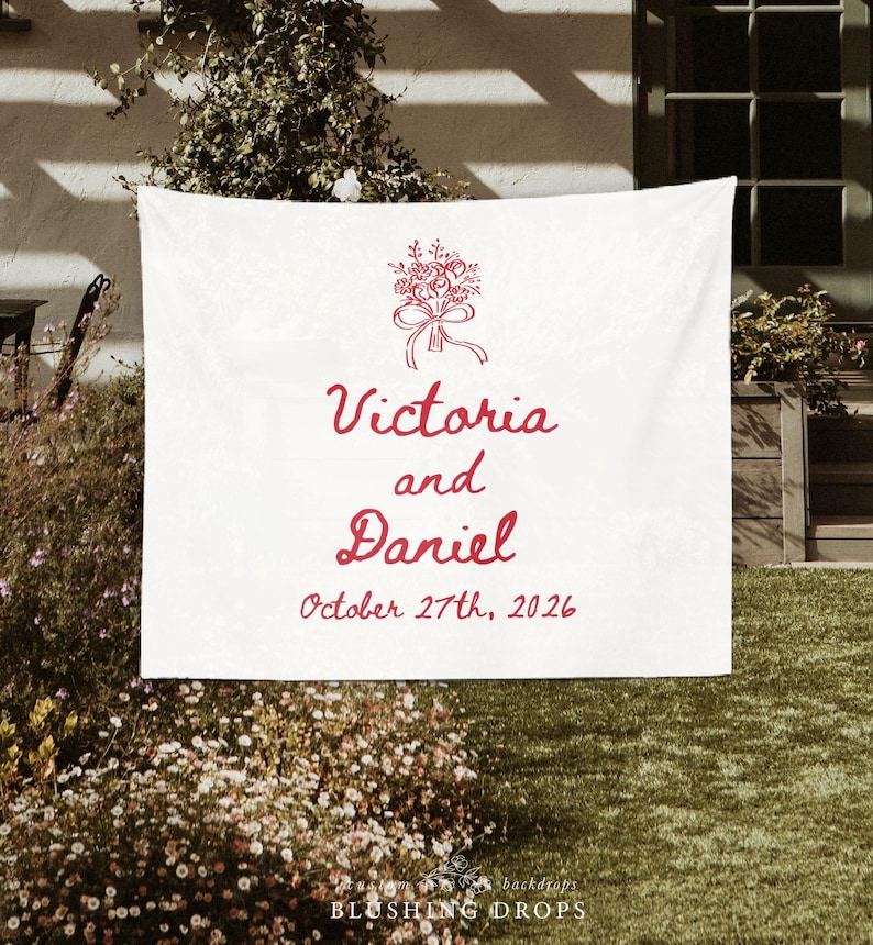 May include: White wedding backdrop with red floral bouquet illustration, the names "Victoria and Daniel", and the date "October 27th, 2026". The backdrop is hanging outdoors with greenery in the background.