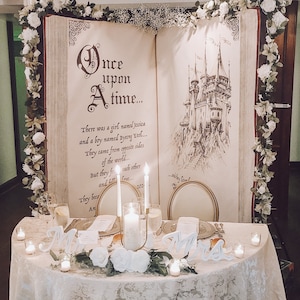 Puede incluir: Una mesa de boda con un mantel blanco, flores blancas y velas. El tel&oacute;n de fondo es un gran libro abierto con el texto "&Eacute;rase una vez..." y un dibujo de un castillo.